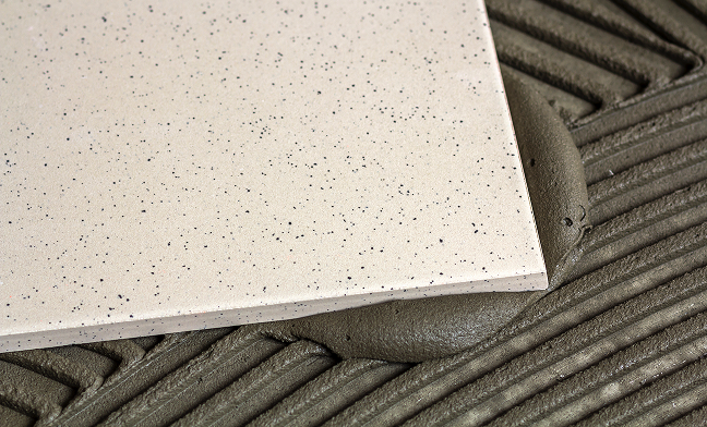 Eine helle Keramikfliese mit dunklen Sprenkeln wird in ein Bett aus frischem, geriffeltem Fliesenkleber gedrückt, wobei etwas Kleber hervorquillt.