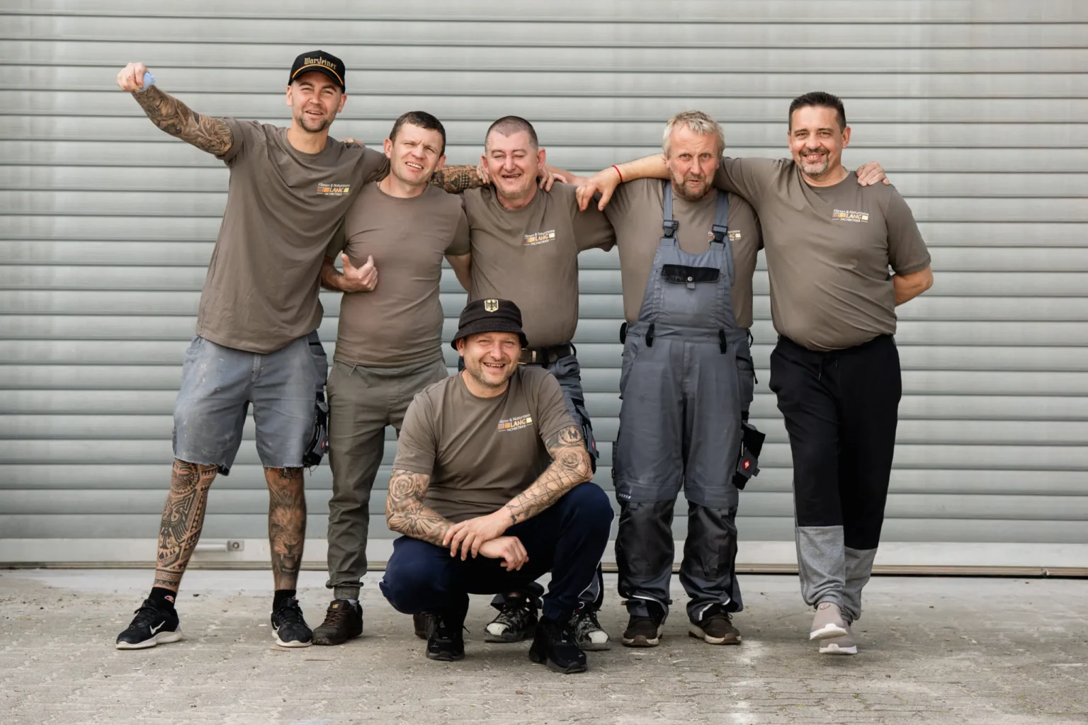 Eine Gruppe von sechs lächelnden Männern in Arbeitskleidung, einige tätowiert, posiert gemeinsam vor einer grauen Wellblechwand.